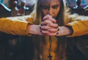 Pessoa em igreja, com as mãos unidas em oração e a cabeça baixa, em um ambiente de luz suave e silêncio.