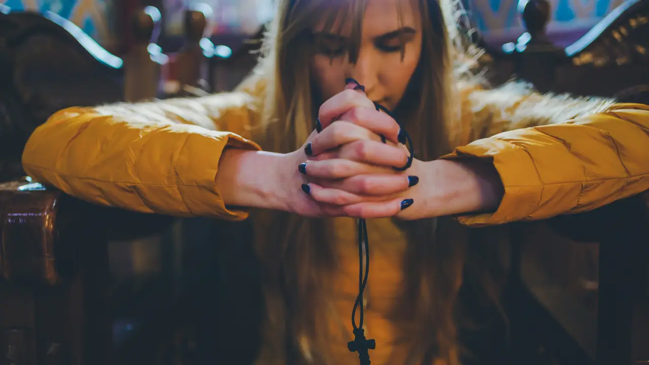 Pessoa em igreja, com as mãos unidas em oração e a cabeça baixa, em um ambiente de luz suave e silêncio.